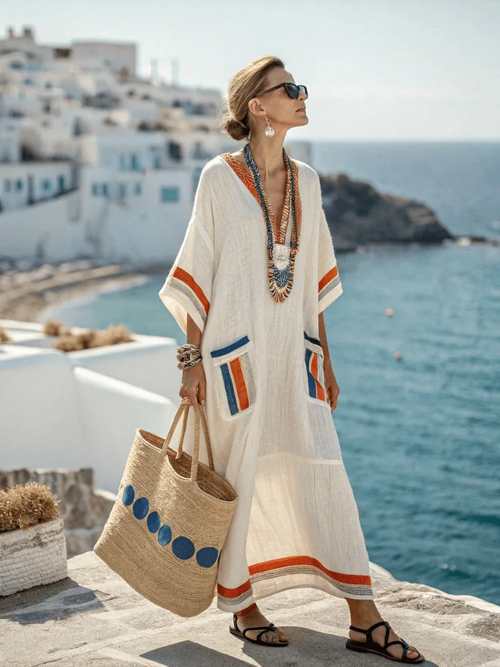 Woman in a white dress with orange and blue patterns, holding a straw bag, standing on a balcony with a scenic view of the sea and buildings.
