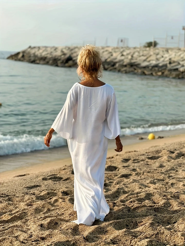 Person in a white dress standing on a beach with ocean and rocks in the background