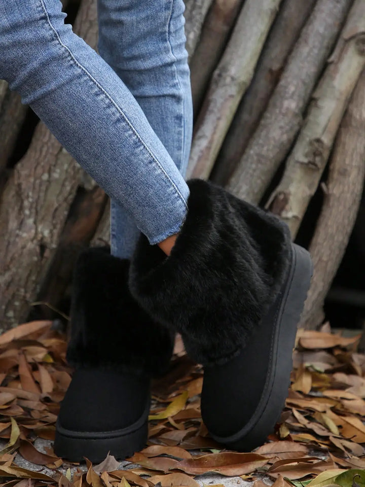 Black faux-fur ankle boots with platform soles, worn with blue jeans on a ground covered in fallen leaves in front of a pile of logs.