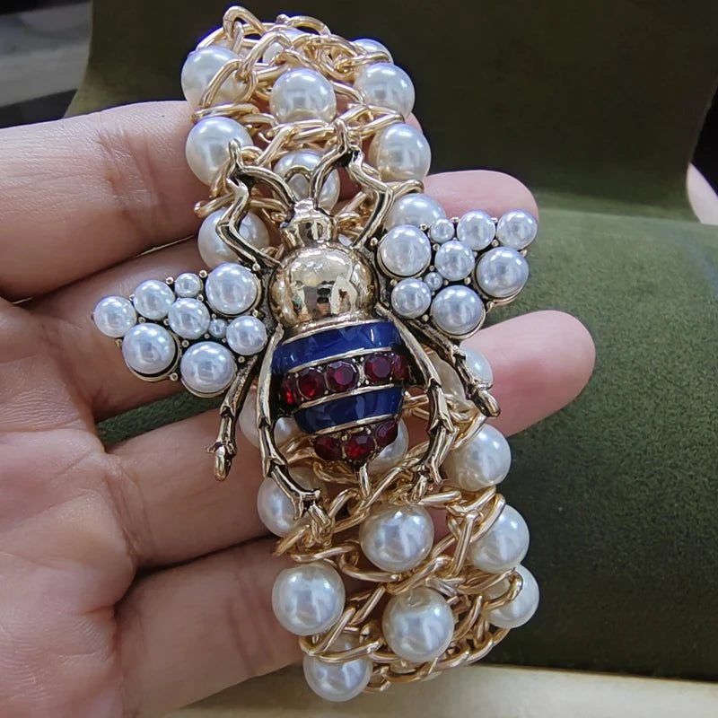 Gold bee-shaped brooch with pearls and red stones held in a hand against a green background