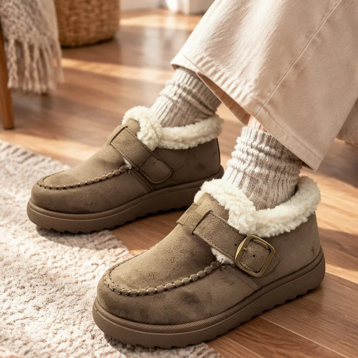 A person wearing taupe-brown suede slip-on moccasin-style slippers with white sherpa lining and a small gold-tone buckle, paired with cream socks and beige pants, standing on a wooden floor.