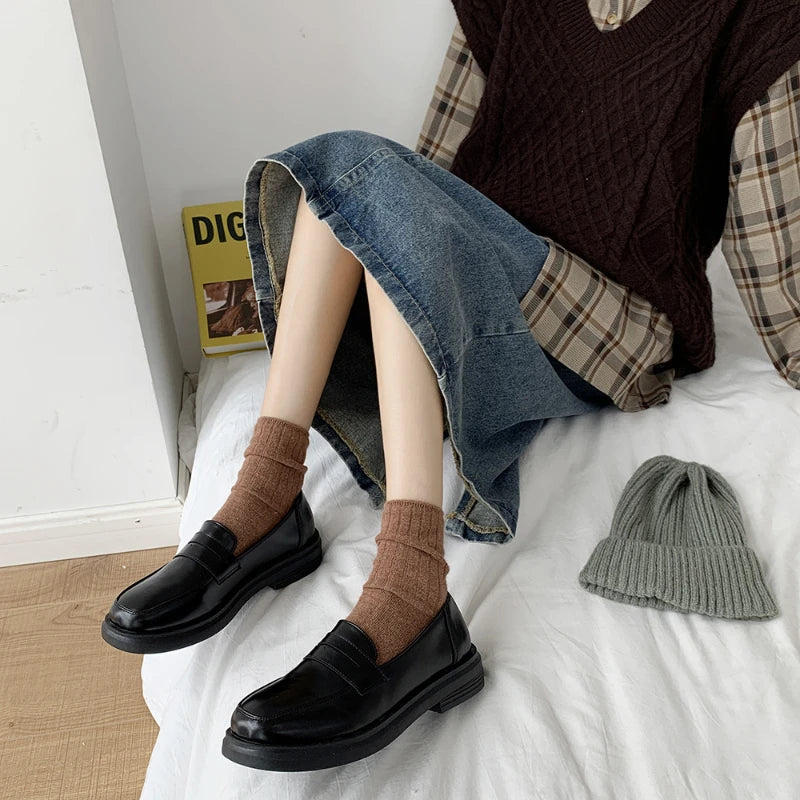 Person wearing black shoes and brown socks sitting on a bed with a hat and magazine in the background.