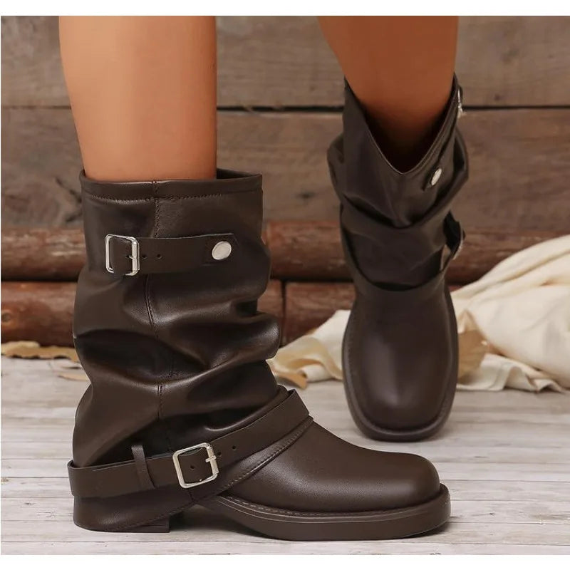 Dark brown slouchy ankle boots with buckle straps, shown on a model's feet against a rustic wood background.