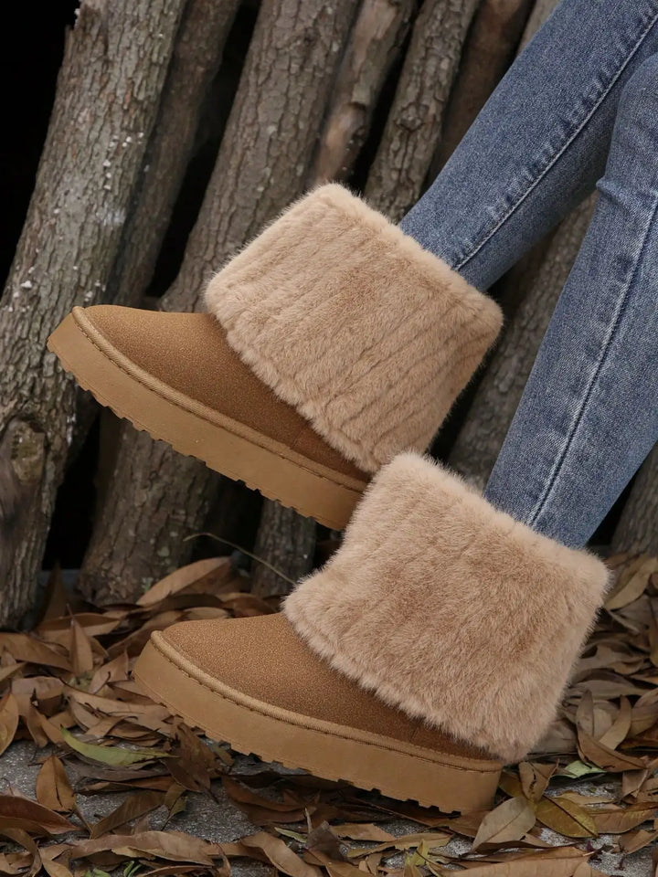 A person wearing tan suede-like ankle boots with thick cream faux-fur cuffs and light tan soles, paired with blue jeans, standing outdoors on dry leaves against a backdrop of tree trunks.