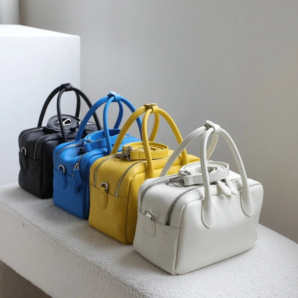 Four structured leather handbags in black, blue, yellow, and white, arranged in a row on a light textured surface against a neutral gray background.