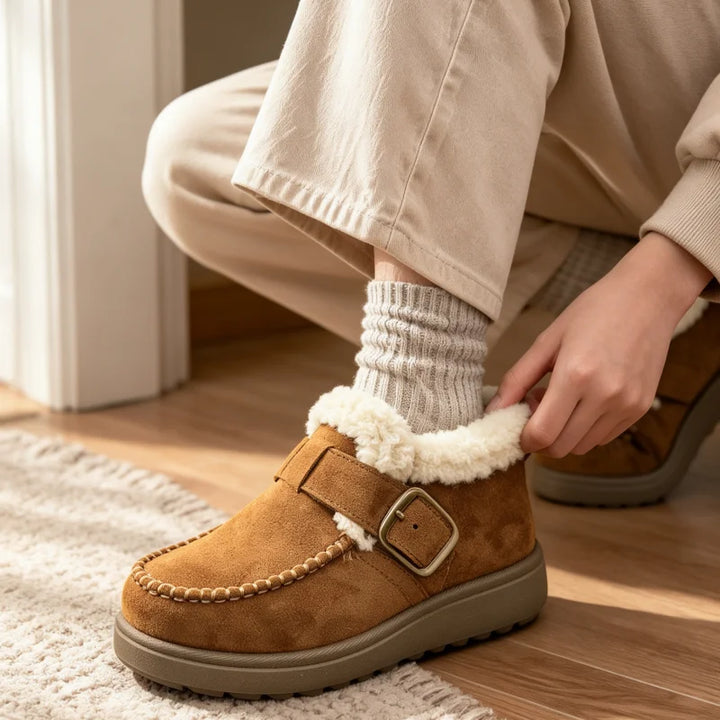 A person wearing a tan suede slip-on moccasin-style bootie with a white sherpa lining and a buckle strap, shown on a wooden floor in a cozy indoor setting.