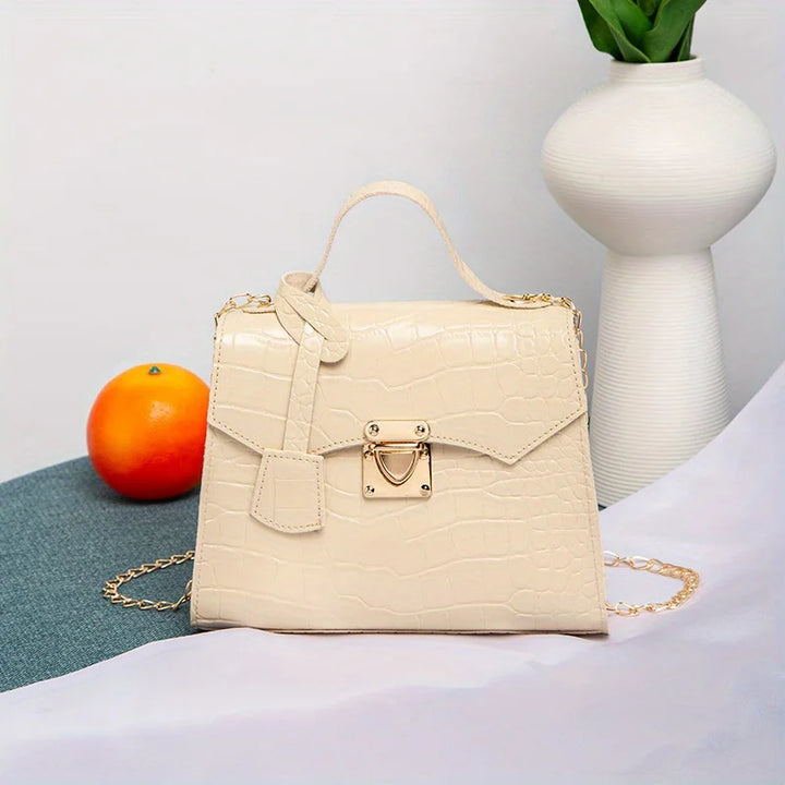 Beige handbag with gold accents on a surface with an orange and vase in the background