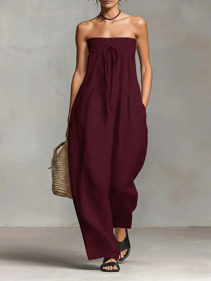 A woman models a strapless, wide-leg jumpsuit in deep burgundy. The jumpsuit features a gathered smocked bodice with a front tie detail and side pockets.