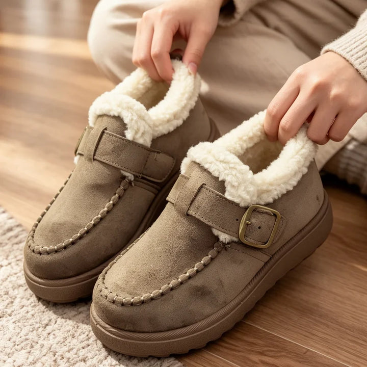 A pair of taupe faux-suede slip-on moccasin-style winter boots with a thick cream sherpa lining, shown being worn with a person's hands pulling the collar up. The boots feature a decorative braided trim along the toe and a gold-tone buckle strap.