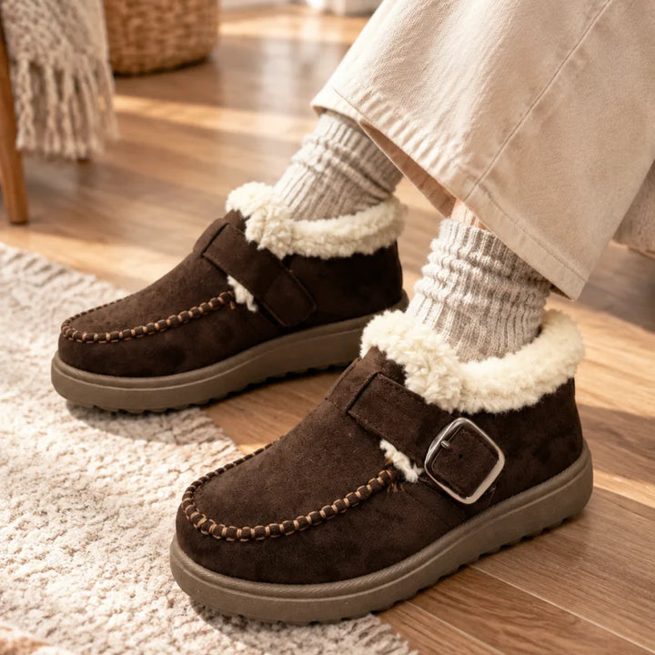 A person wearing dark brown suede slipper-style shoes with cream sherpa lining and a buckle strap, paired with beige pants and ribbed socks, standing on a wooden floor.