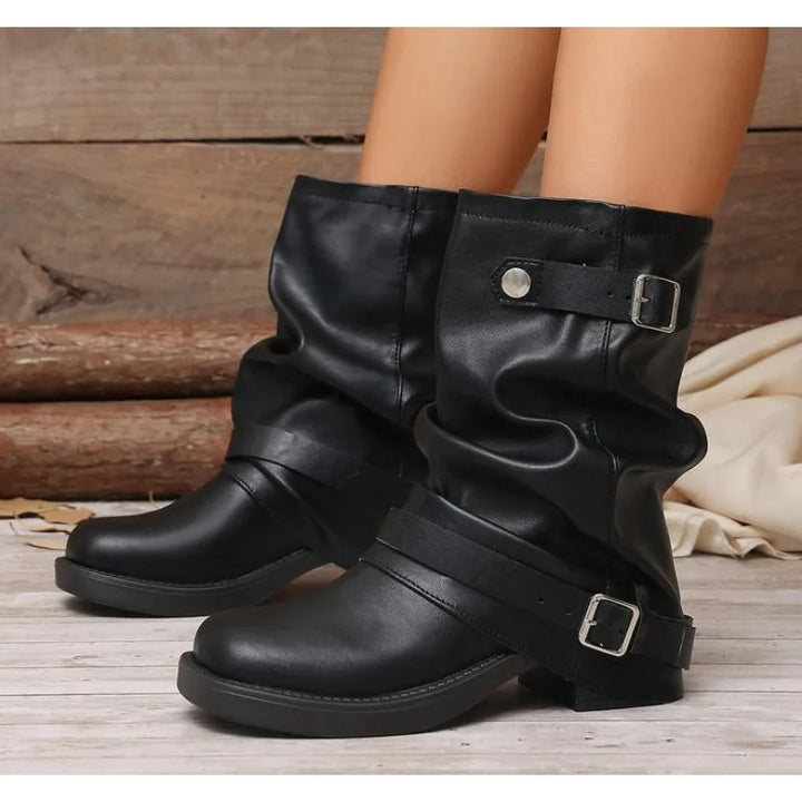 A pair of black faux-leather slouchy mid-calf boots with buckle straps, shown on a model's feet against a rustic wood background.