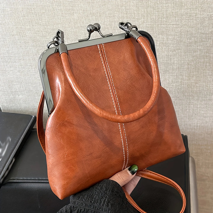 Brown leather handbag with a metal frame held by a hand against a neutral background