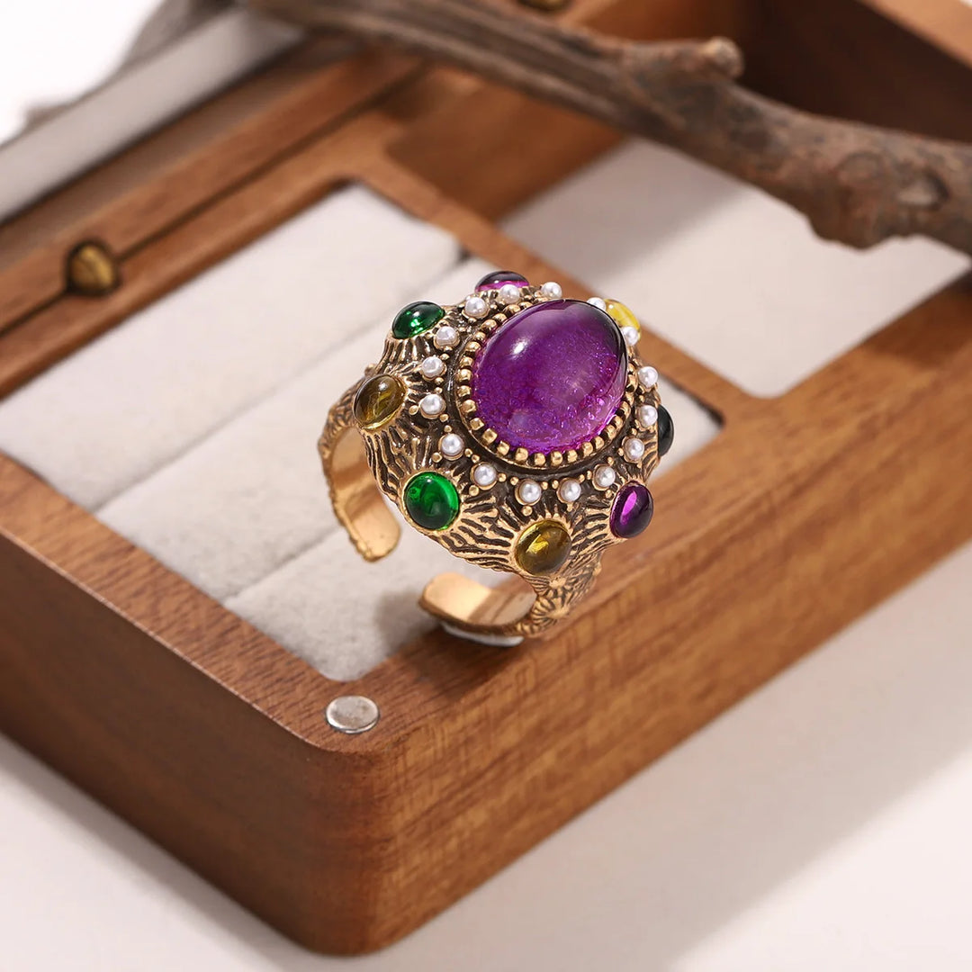 Gold statement ring with oval purple centerpiece, pearl-like stones, and green gem drops