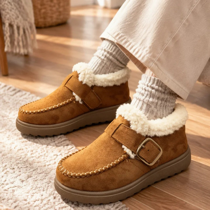 A person wearing tan suede slip-on moccasin-style winter boots with white sherpa lining and a buckle strap, paired with cream socks and light pants, standing on a wooden floor.