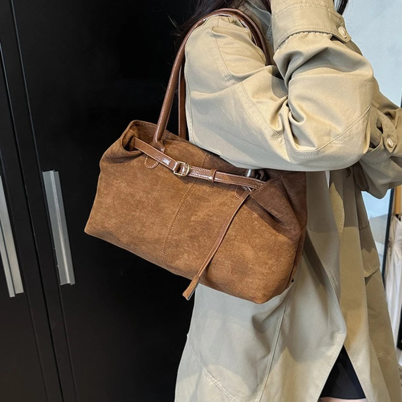 Women’s suede tote bag in brown with top handle, buckle strap, and hanging tassel.