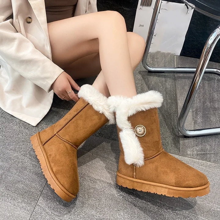 A person wearing tan suede-like mid-calf winter boots with white faux-fur trim and a decorative button detail, seated indoors on a gray floor.