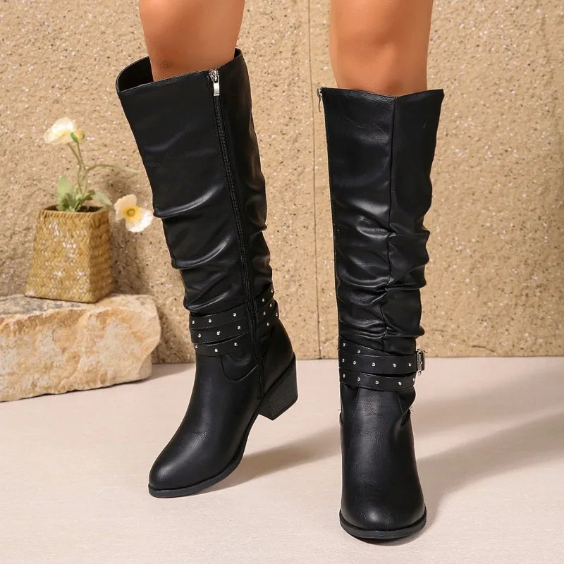 A pair of black knee-high slouchy boots with studded straps and low block heels, shown on a model's legs against a textured beige wall and stone floor.