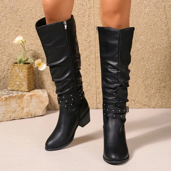A pair of black knee-high slouchy boots with studded straps and low block heels, shown on a model's legs against a textured beige wall and stone floor.