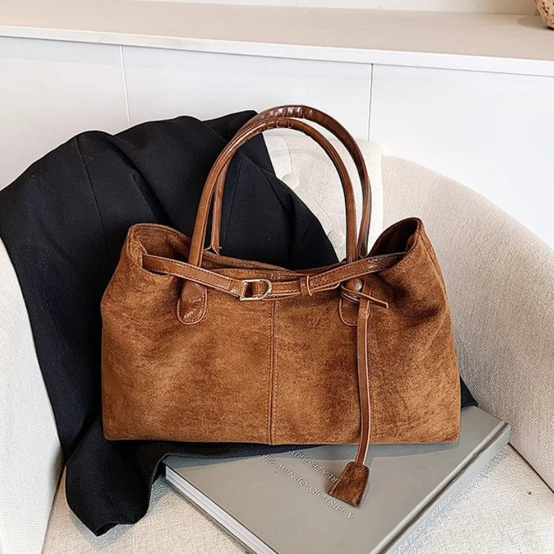 Women’s suede tote bag in brown with top handles, hanging tassel, and side seam panel.