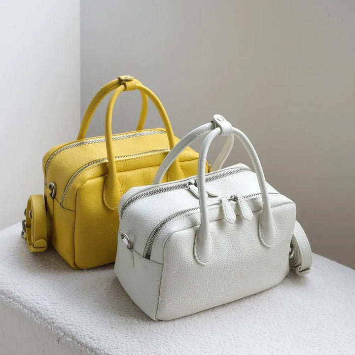 Two structured pebbled leather handbags with double top handles and zip-top closures, shown on a white textured surface against a light gray wall. The bag in the back is mustard yellow, and the bag in the front is off-white/cream.