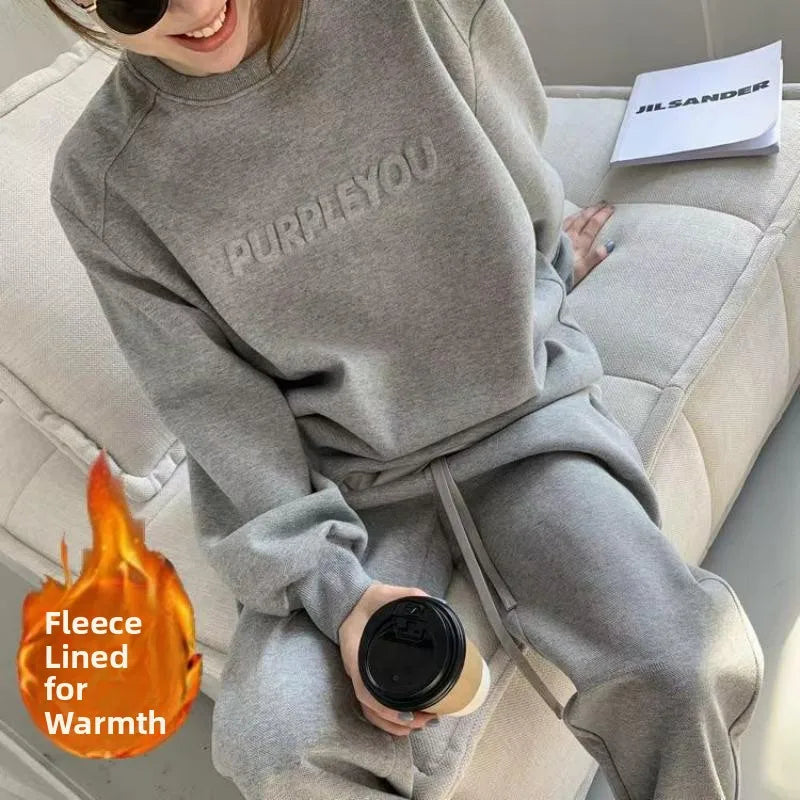 A person wearing a light gray fleece sweatshirt and matching sweatpants, sitting on a light-colored couch and holding a coffee cup. The sweatshirt has 'PURPLEYOU' printed across the chest.
