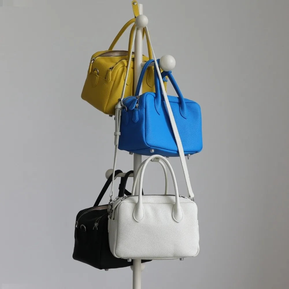 Four structured handbags in yellow, blue, black, and white hang from a white display stand against a light gray studio background.