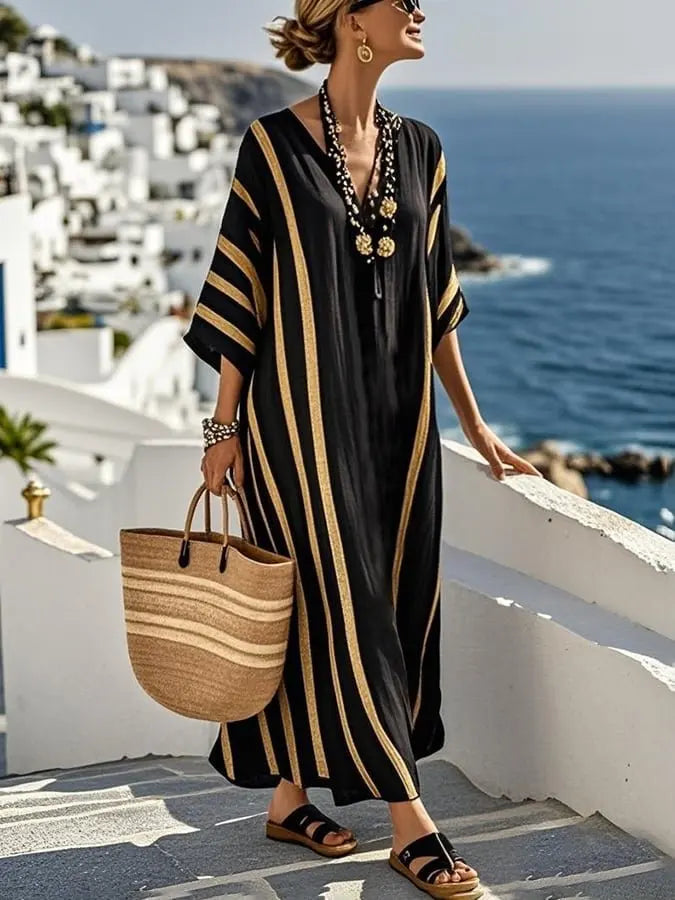 Woman in a black and gold dress with a straw bag standing on a balcony overlooking the ocean and white buildings.