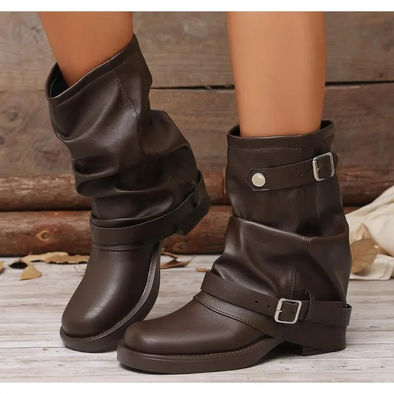 Dark brown faux leather slouchy ankle boots with buckle strap details, shown on a model's feet against a rustic wood background.