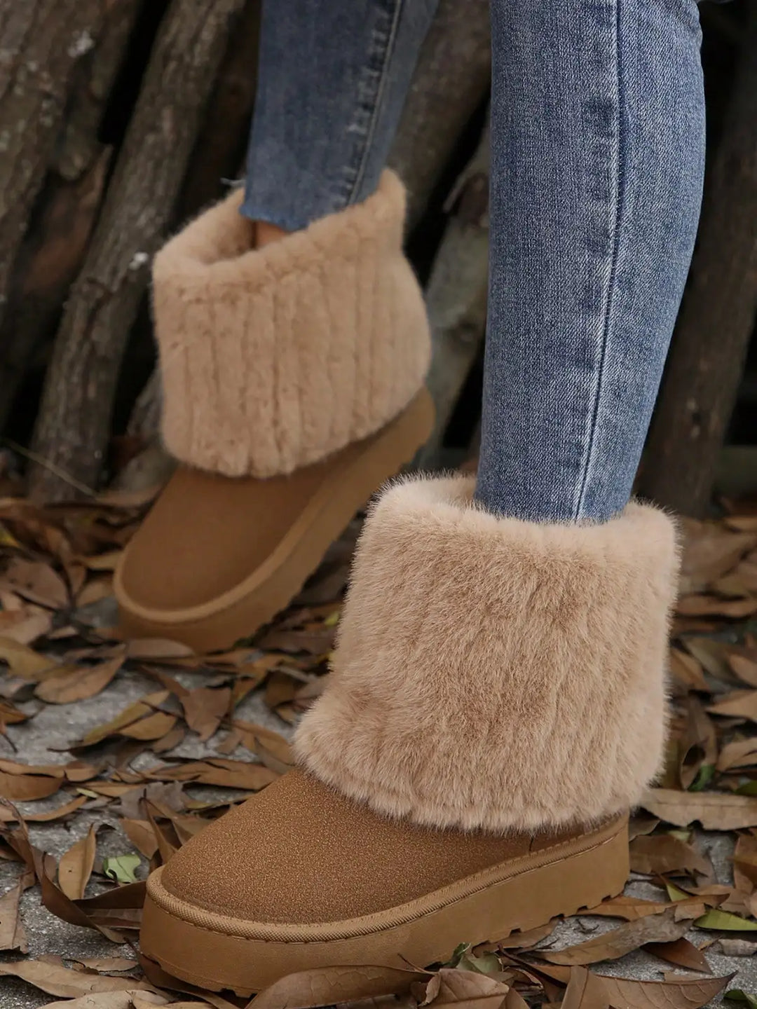 A person wearing tan suede-like ankle boots with fluffy cream faux-fur trim, paired with blue jeans, standing outdoors on a ground covered in dry leaves.