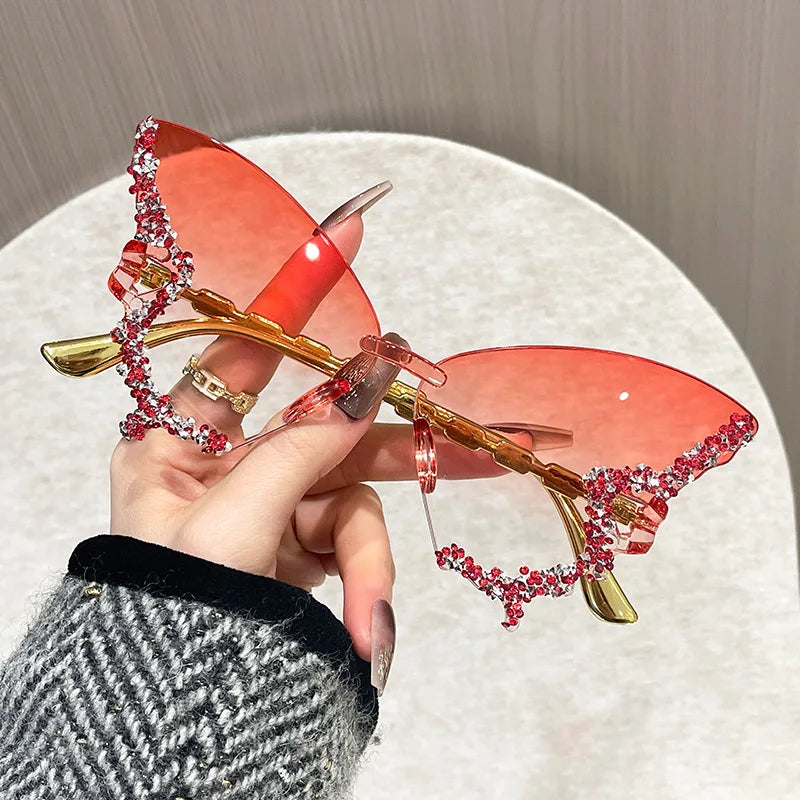 Butterfly-shaped sunglasses with red lenses and gold frames held by a hand.