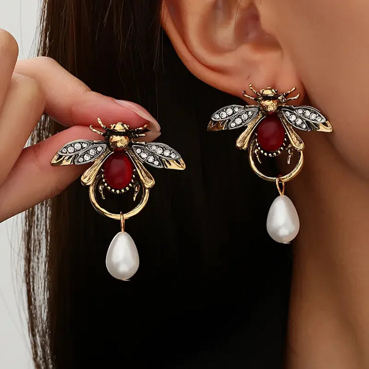 Bee-shaped earrings with red gemstones and pearl drops on a reflective surface.