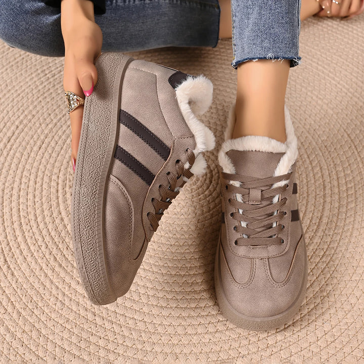 A person wearing taupe suede low-top sneakers with white faux-fur lining and black side stripes, shown from the knees down while sitting on a beige woven rug.