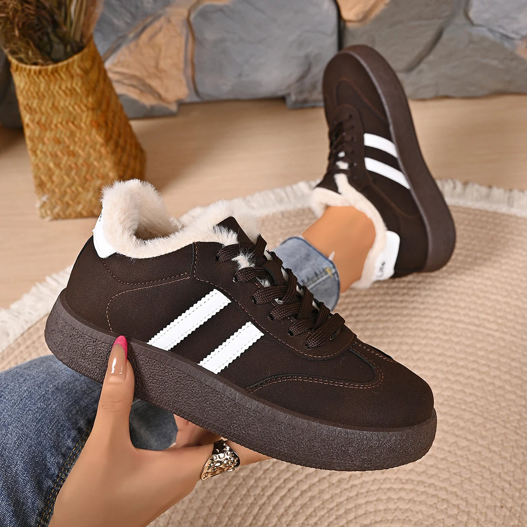 A person holds up a dark brown low-top sneaker with white side stripes and a white faux-fur collar, while wearing the same shoe on one foot in a cozy indoor setting.
