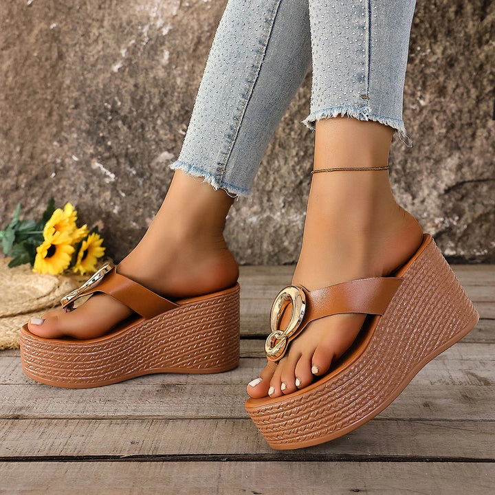 Brown platform sandals with gold accents worn by a person on a wooden surface with a stone wall background.
