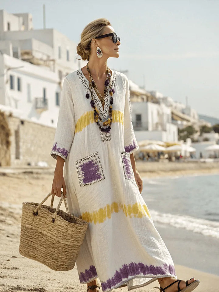 Woman in a white dress with purple and yellow patterns walking on a beach.