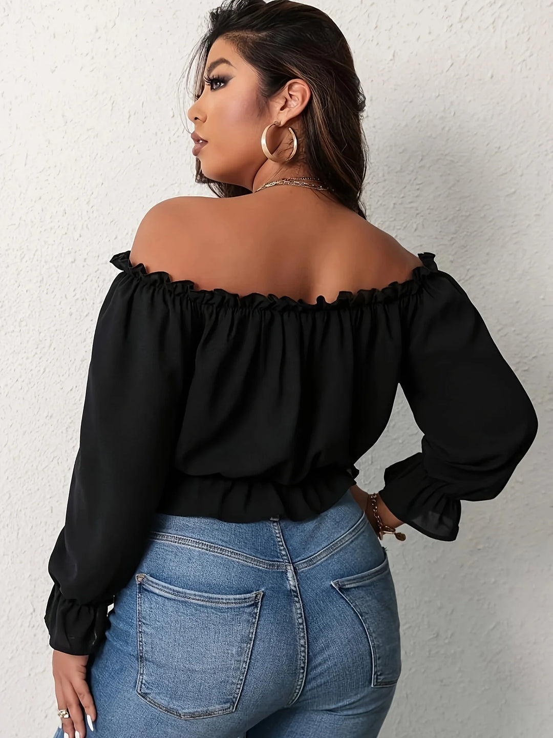 Woman wearing a black off-shoulder top and blue jeans against a white wall