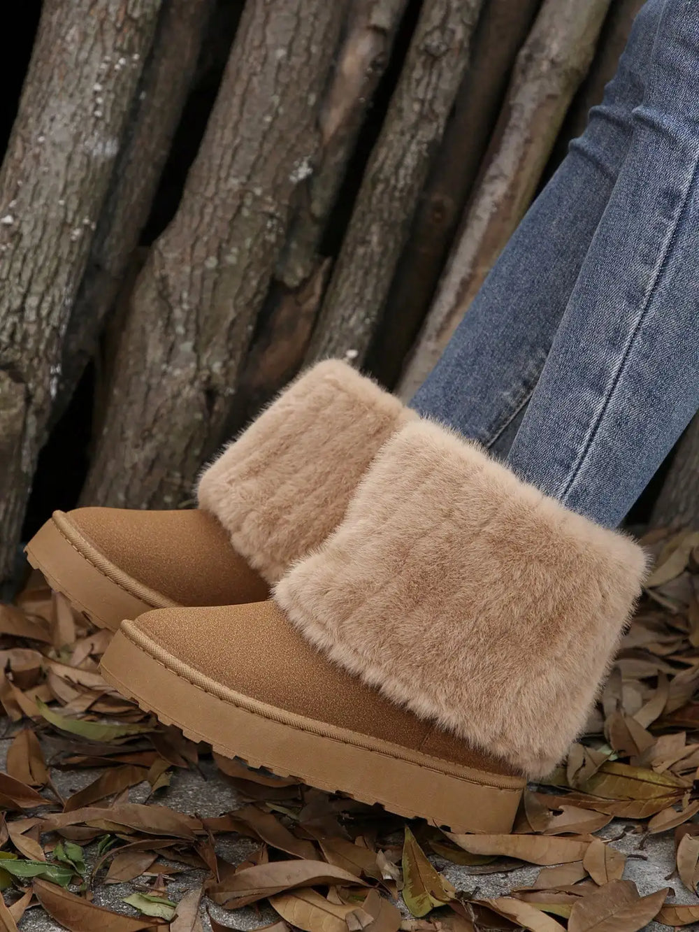 A pair of tan suede-like slip-on boots with fluffy beige faux-fur trim, shown worn with blue jeans on a ground covered in dry leaves in front of a wood pile.