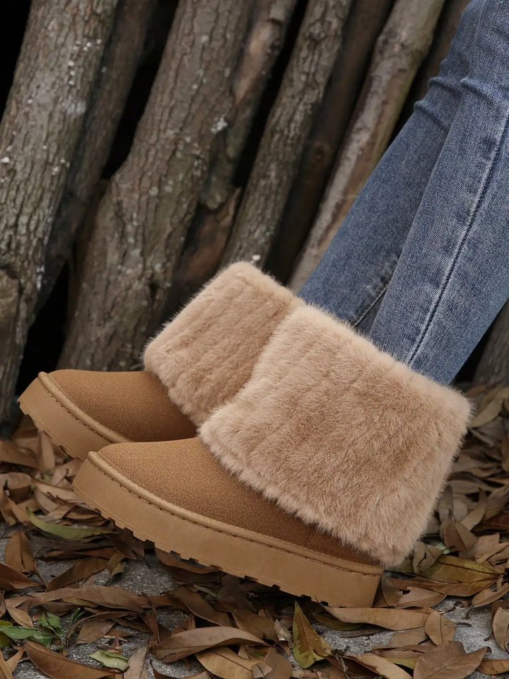 A pair of tan suede-like slip-on boots with fluffy beige faux-fur trim, shown worn with blue jeans on a ground covered in dry leaves in front of a wood pile.