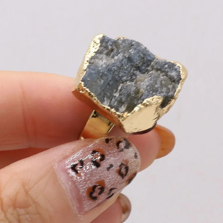 Gold ring with a rough stone held by a hand with patterned nails on a light background