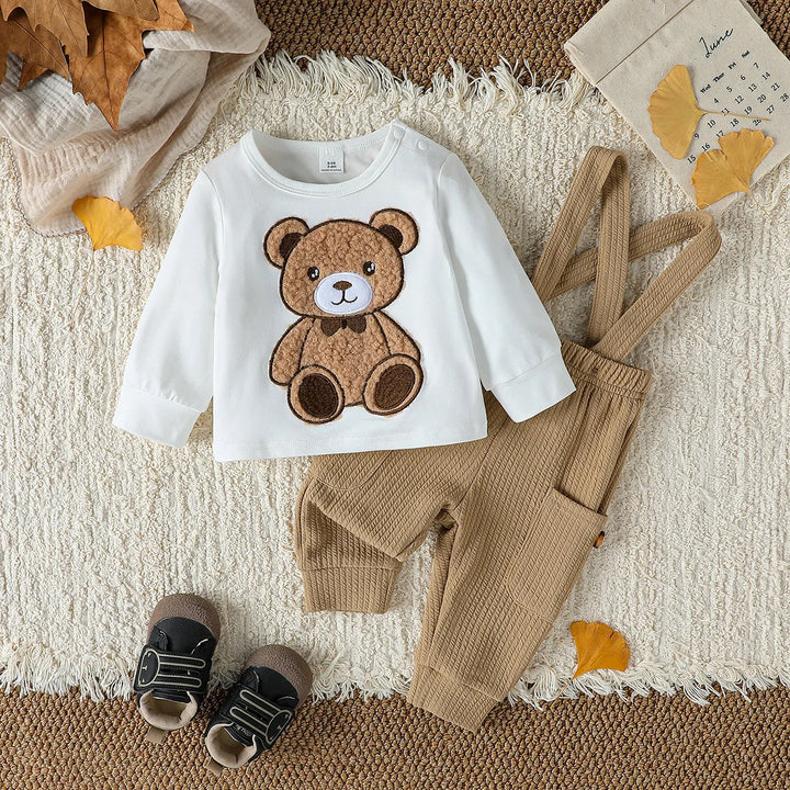 Children's outfit with white shirt featuring a teddy bear, beige pants, and shoes on a textured surface.