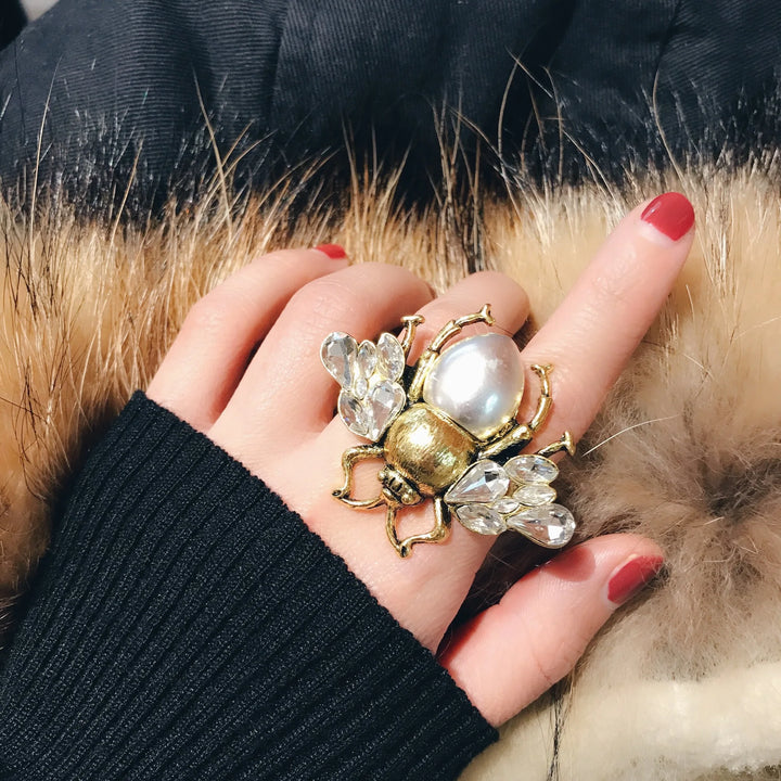 Hand wearing a decorative ring with pearls and crystals, against a fur background.