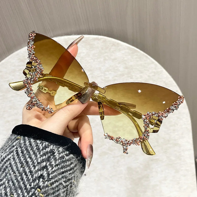 Gold butterfly sunglasses with gemstones held by a hand against a neutral background