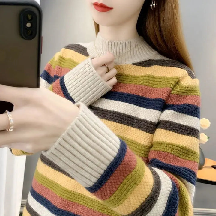 A woman wearing a chunky knit mock-neck sweater with horizontal stripes in beige, yellow, green, navy, and dusty pink, holding a dark smartphone in a mirror selfie.