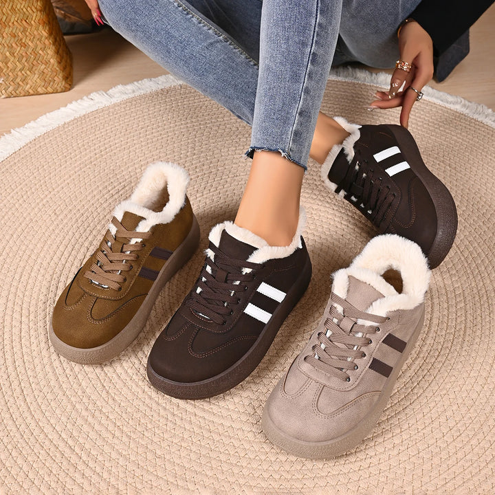 Four low-top winter sneakers with thick white faux-fur lining are arranged on a round woven rug, with a person's legs and hands visible in jeans holding one shoe. The shoes come in brown, dark brown, taupe, and olive tones with dark side stripes and white stripe accents.