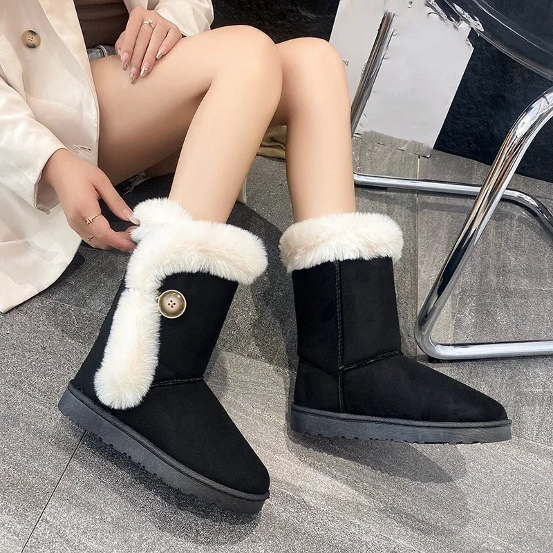 A person seated indoors wearing black mid-calf boots with thick white faux-fur trim and a decorative gold-tone button. The photo is cropped to the lower torso, legs, and boots, with a chair and gray floor visible in the background.