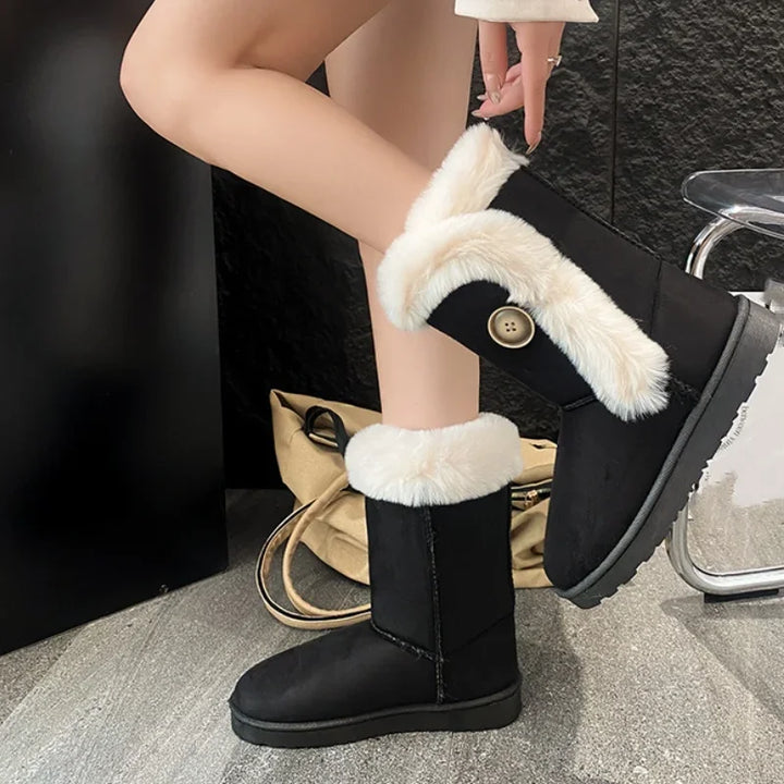 A person wearing black mid-calf winter boots with thick white faux-fur trim and a decorative gold-tone button, shown in a styled indoor setting.