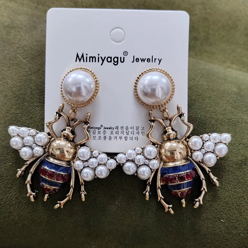Bee-shaped earrings with pearl details on a Mimiyagu Jewelry card against a green background