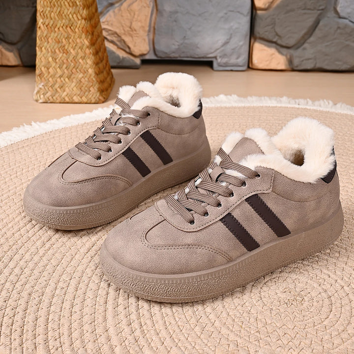 A pair of taupe-brown low-top winter sneakers with white faux-fur lining and black side stripes, displayed on a round woven rug.
