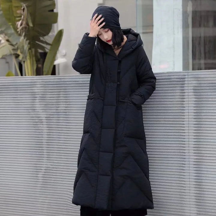 Person wearing a long black puffer coat standing against a light-colored wall.