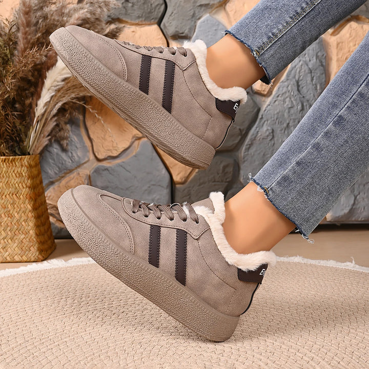 A pair of taupe suede low-top sneakers with white faux-fur lining and three dark side stripes, shown being worn with blue jeans in a cozy indoor setting with a stone wall and woven rug.